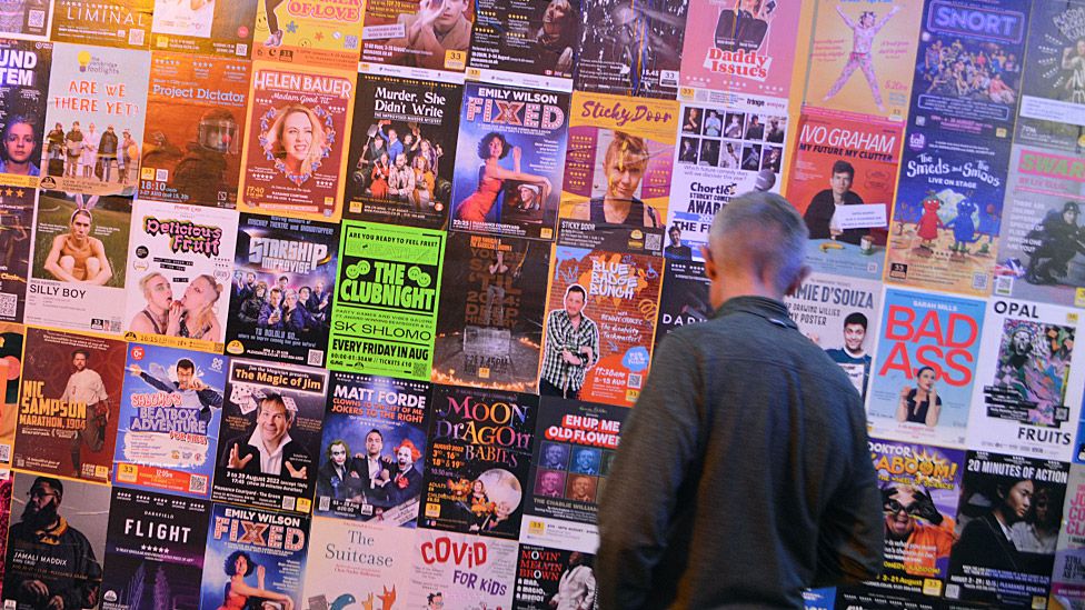 A man standing in front of a wall of posters for shows at the Edinburgh Fringe
