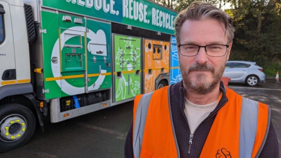 Councillor Chris Watts looks into the camera wearing a hi-vis jacket in front of a recycling lorry