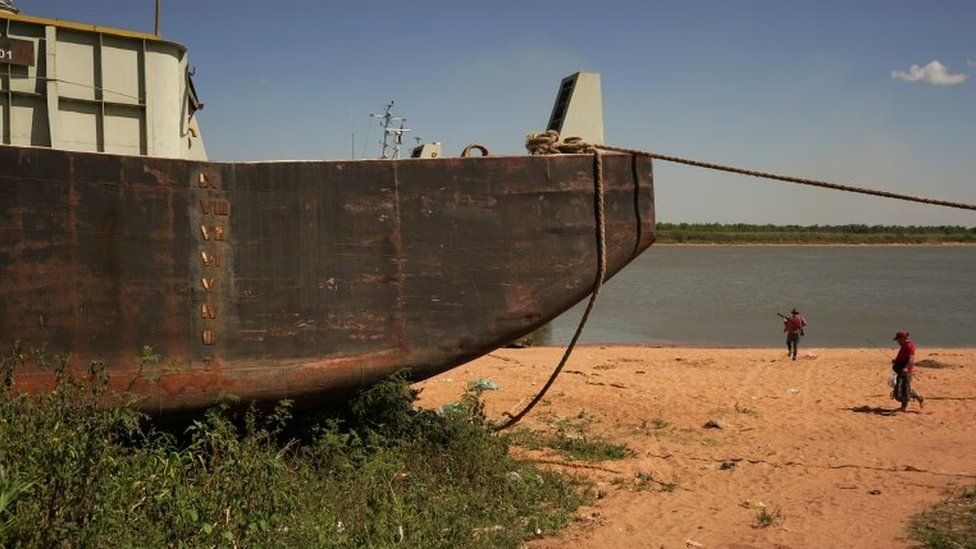 People walk past a stranded barge on the shore of the Rio Paraguay (Paraguay River), which flows down to the Parana River, as the lack of rain in Brazil, where the river originates, has brought water levels down, forcing cargo ships to reduce the amount of grains that are loaded for export, in Ypane, Paraguay August 30, 2021