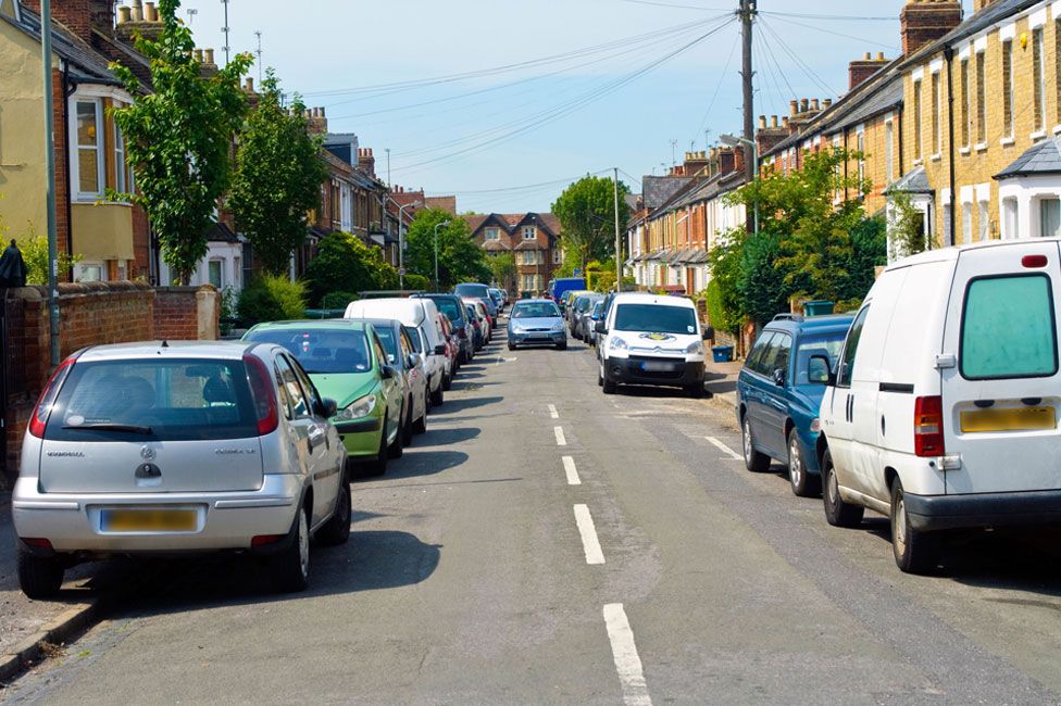 Cars parked on both sides of road, on pavements, with a car coming down the road