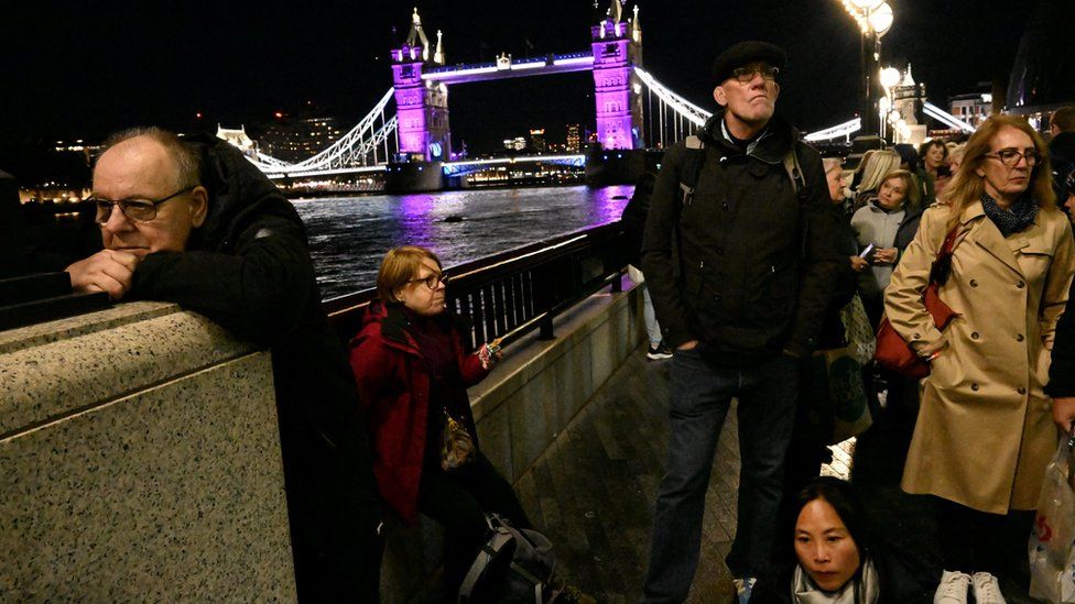 People wait in a queue to pay their respects as the Tower Bridge is lit up in purple