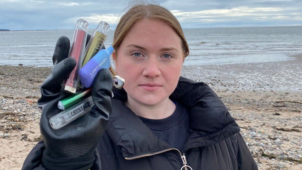 A woman holding a few vapes in front of a beach