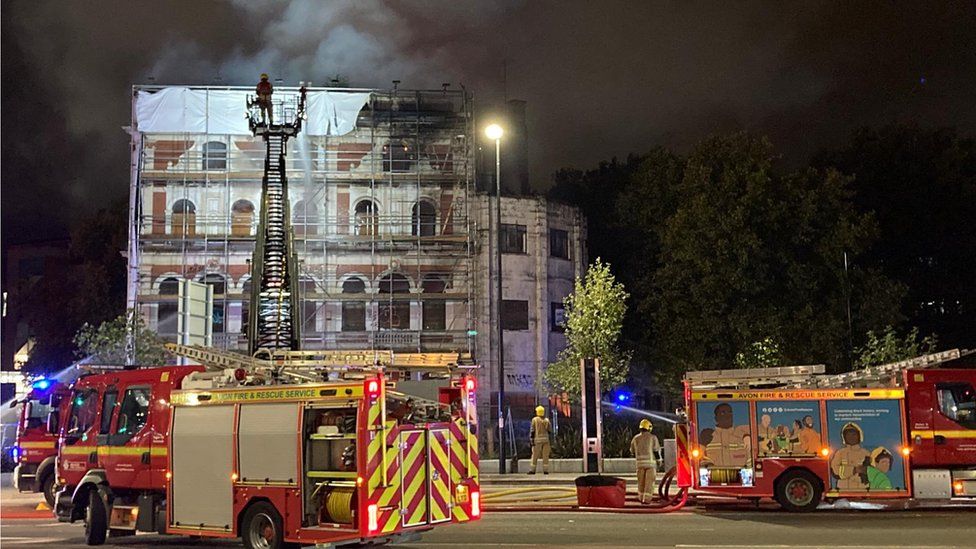 Burning Grosvenor Hotel and fire engines in Bristol. Although the image was taken at night, smoke can be seen rising from the building. Two fire engines are in the foreground of the shot, with a number of crew members stood nearby.