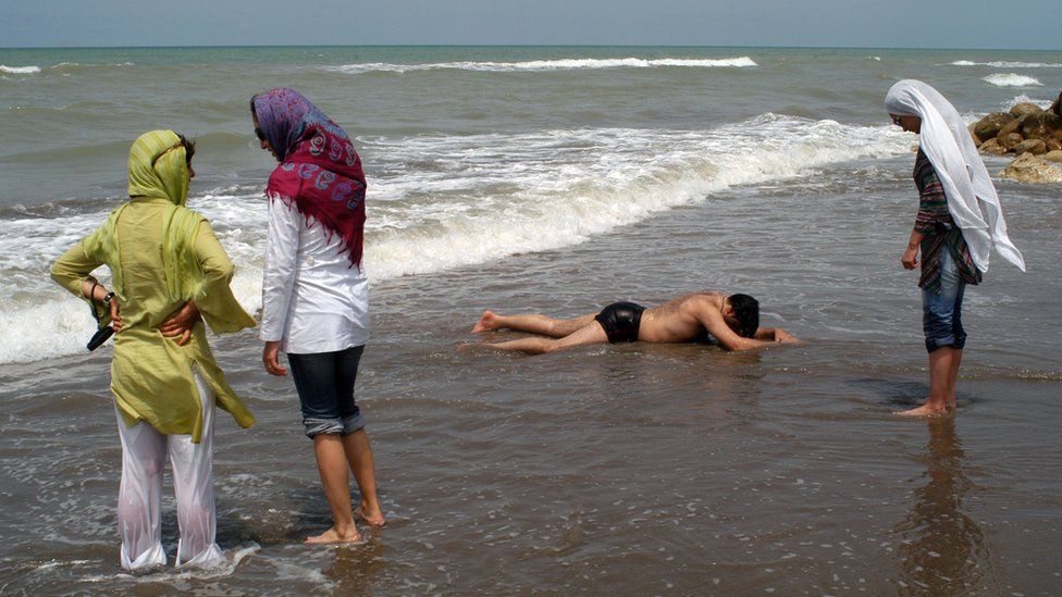 Women paddle in the sea fully-clothed while a man in swimming pants rests on the sand on 10 July 2005