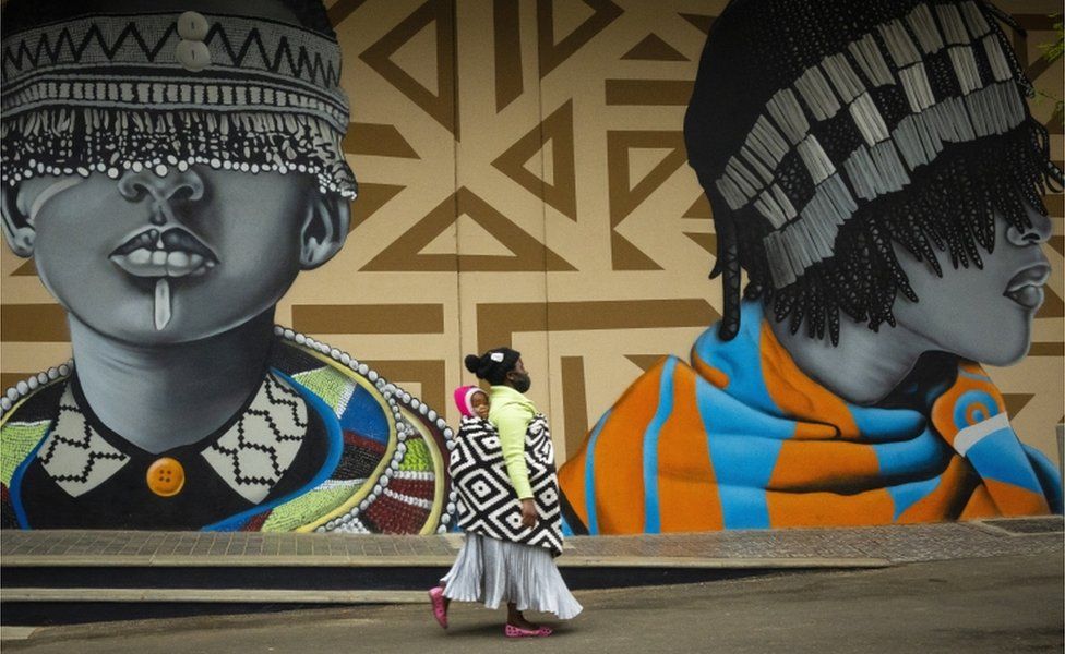 A mother carries her child on her back as she walks past murals featuring graphic prints and two elaborately dressed figures.