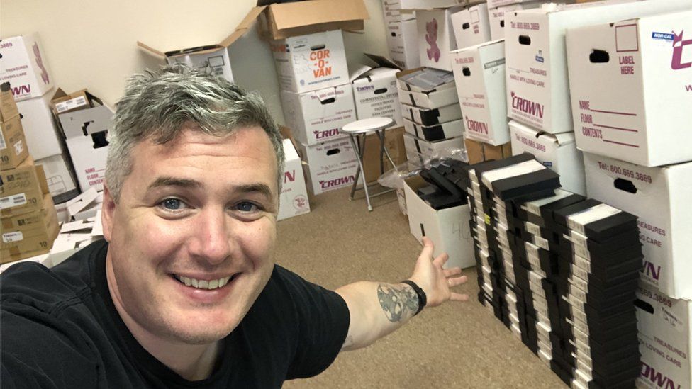 Danny O'Dwyer is in an office room with white walls and beige carpet. He's surrounded by white cardboard boxes - the style you'd usually use if you were moving house. Most of them have a company name - Crown - printed on them, along with spaces for delivery details. The boxes are in stacks at least three-high, and tapes can be seen poking out the top of some. Danny's in the front of the frame, smiling and gesturing towards the haul behind him.