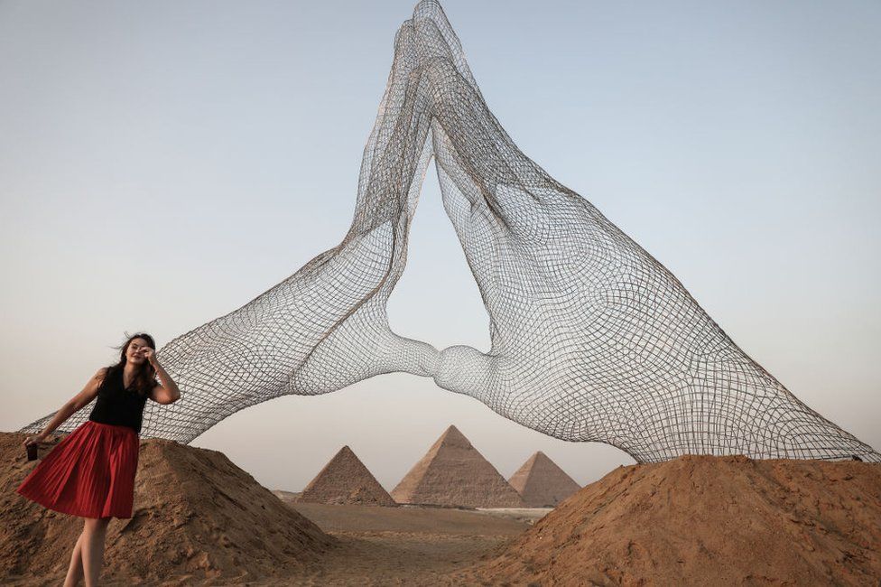 A woman stands next to a large mesh sculpture of human hands.
