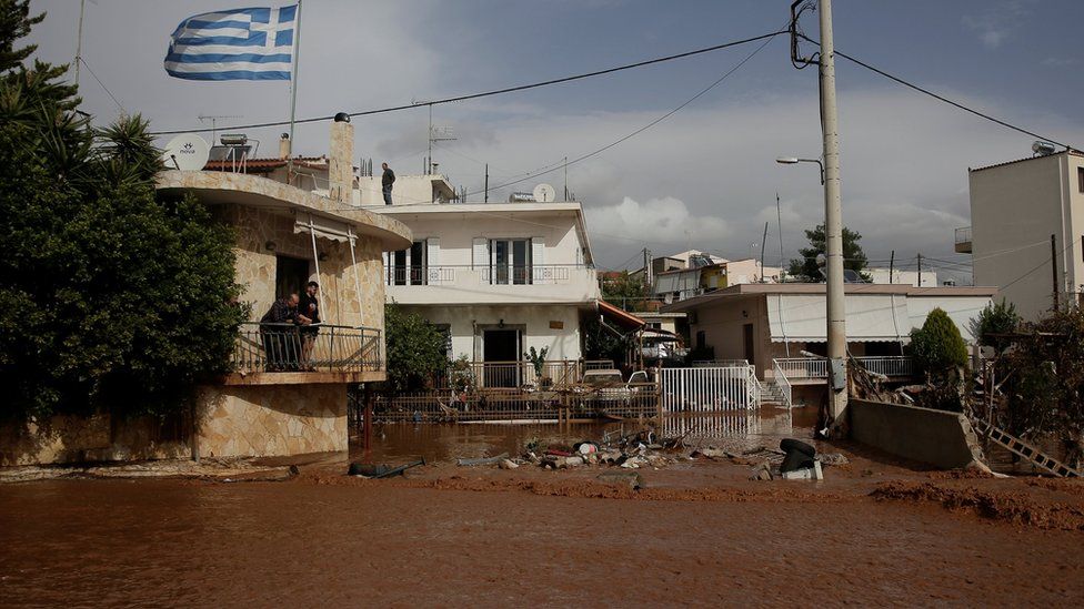 A Greek national flag flutters atop a terrace as locals observe a flooded street following heavy rainfall in the town of Mandra
