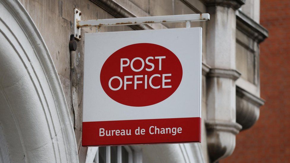 A stock image shows a 'Post Office' sign on the exterior wall of a branch