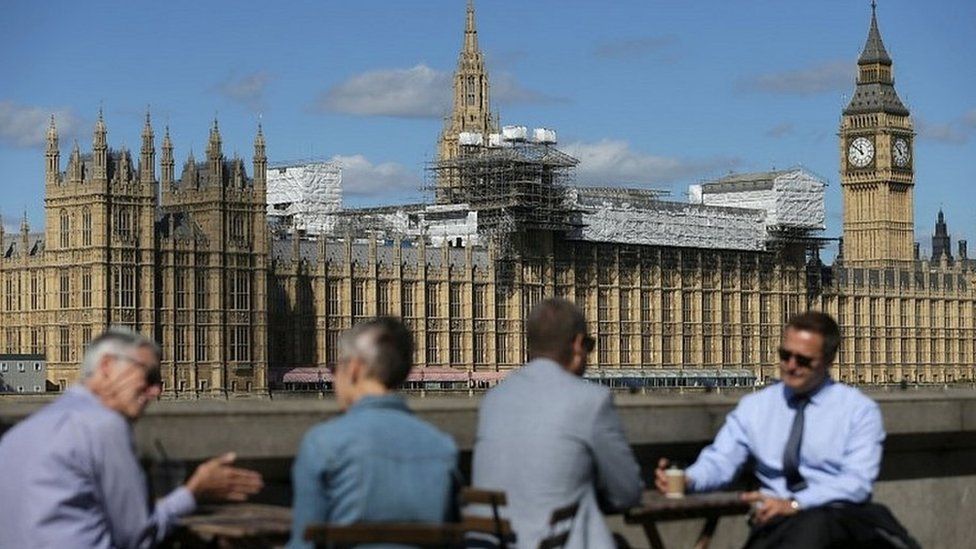 Office workers sit by the Thames, with the Palace of Westminster in the background