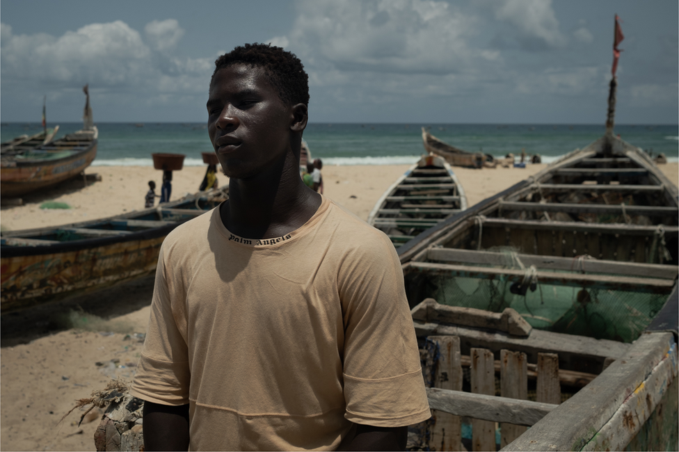 Assane Niang, a 23-year-old fisherman stands on the beach next to pirogue.