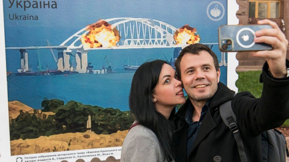 A woman and man pose in front of an artist's impression of the Crimean bridge explosion in Kyiv, Ukraine