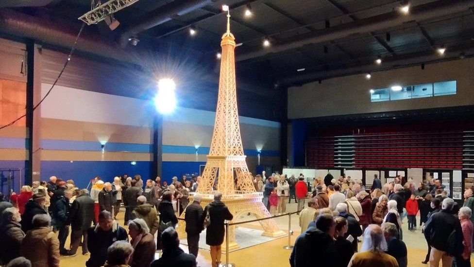 Tall Eiffel Tower made out of matchsticks in the middle of a room full of people
