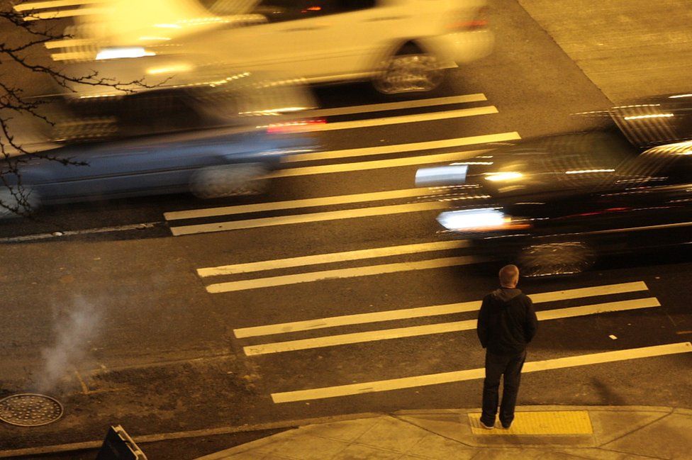 A man waits to cross the road as cars speed past him