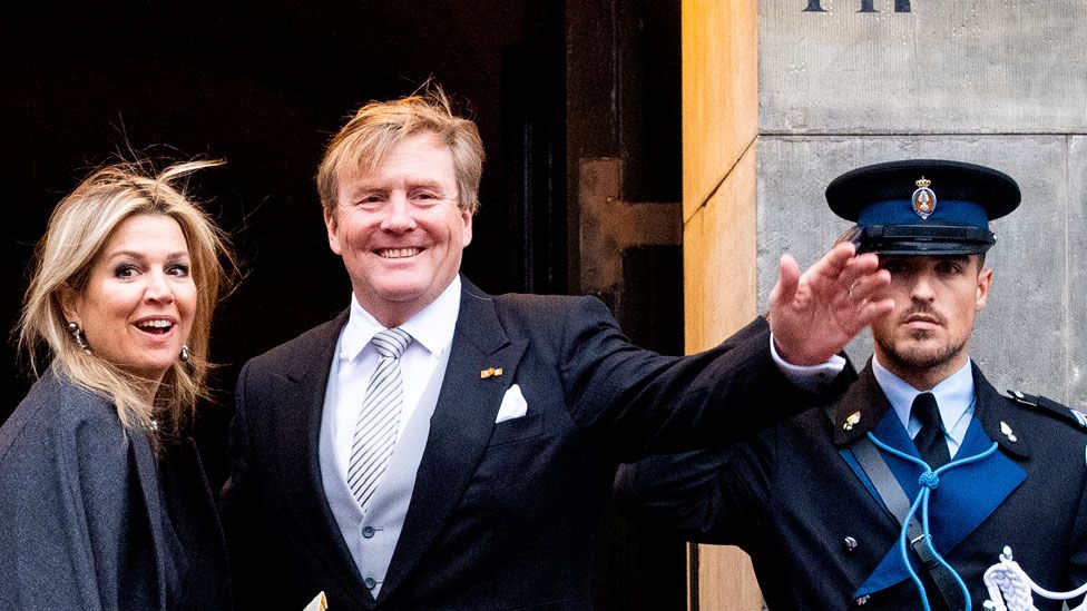 King Willem-Alexander of The Netherlands and Queen Maxima are pictured outside a building waving to the media, while a nearby guard salutes
