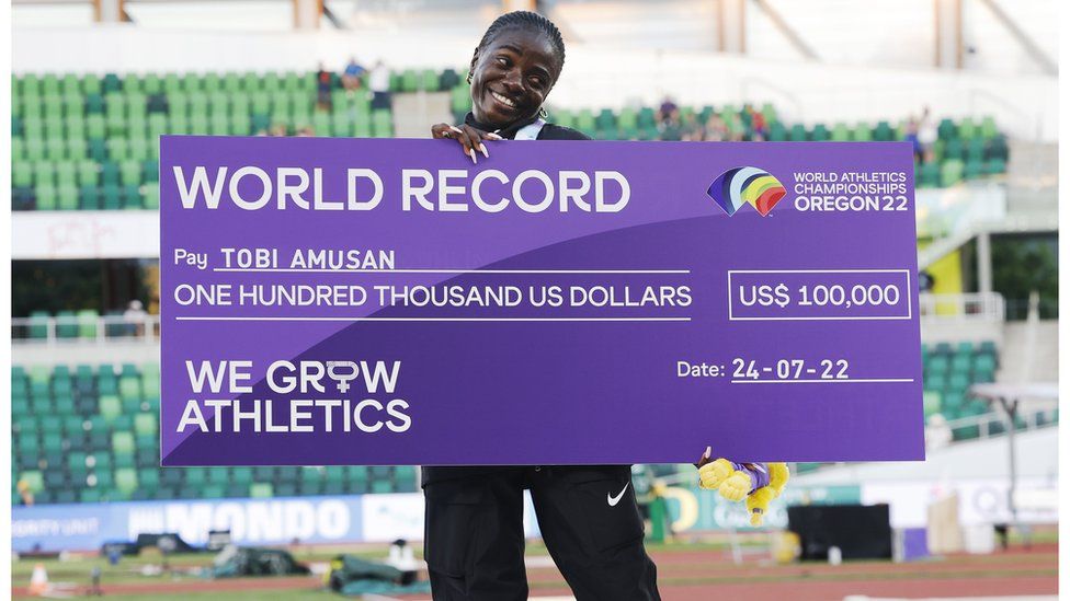 Tobi Amusan holding a giant placard that says World Record on it and the sum she has been paid: $100,000. She is smiling whilst standing on the medals podium on Sunday, in Oregon, the US - Sunday 24 July 2022