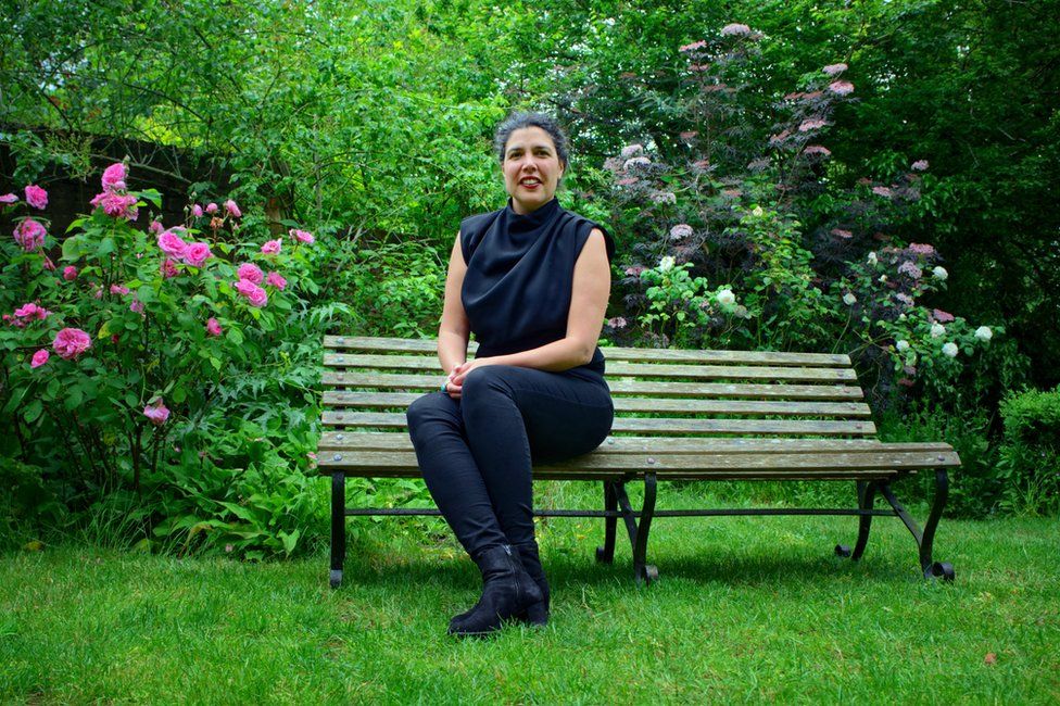 Dr Annouchka Bayley sits on a bench outside surrounded by a pink rose bush to the left and a sambuca nigris to her right. She is wearing black leggings and a sleeveless black high-necked top. She has a quizzical expression