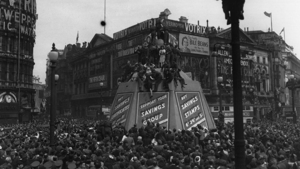 Crowds in Piccadilly Circus climb lampposts and the Eros statue's protective container, on VE Day.