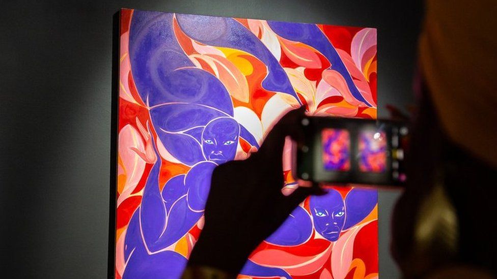 Viewer taking a photo of Tunji Adeniyi's artwork which consists of two painting in blue, red, pink and orange colours.