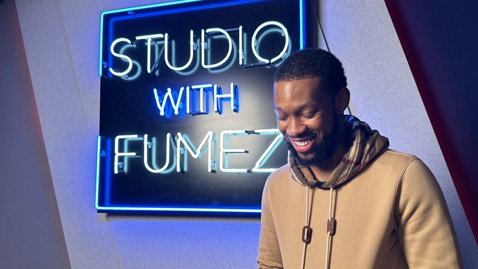 A man wearing a light brown hoodie smiles while looking down, standing in front of a neon sign that reads "Studio with Fumez"