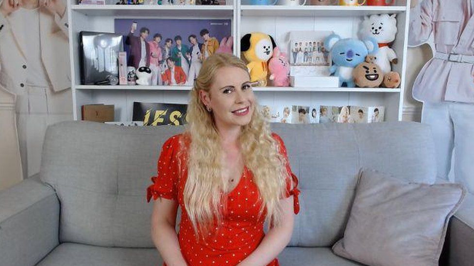 Jenny has long wavy blonde hair. She's wearing a red spotty dress and is sat on a grey sofa. In the background there is a book shelf full of soft cuddly toys and a cardboard cutout of K-Pop boyband.