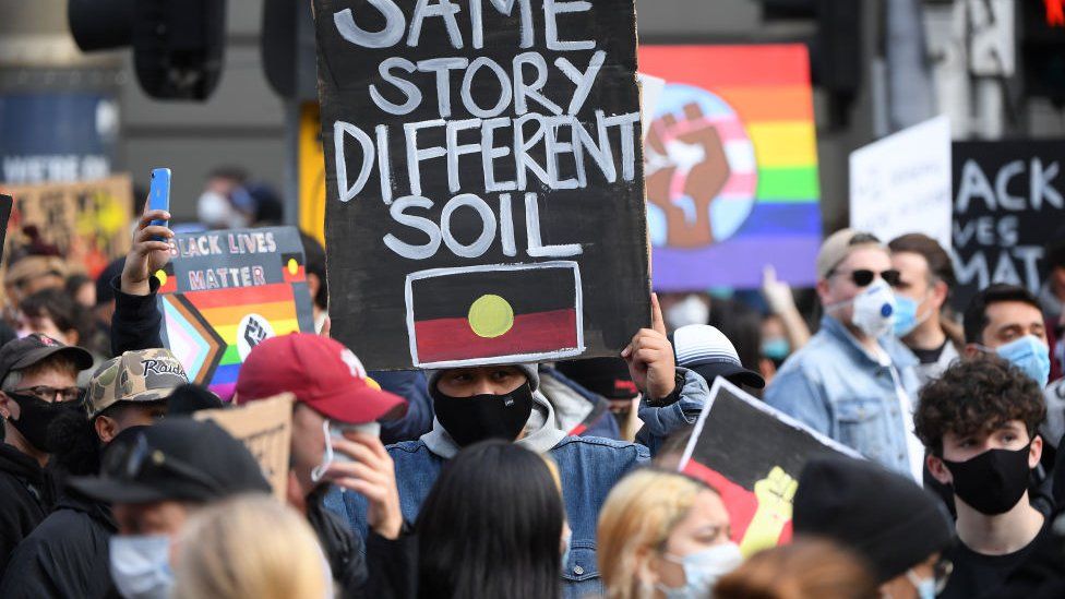 A protests holds a placard with Aboriginal flag which says: "Same story, different soil"
