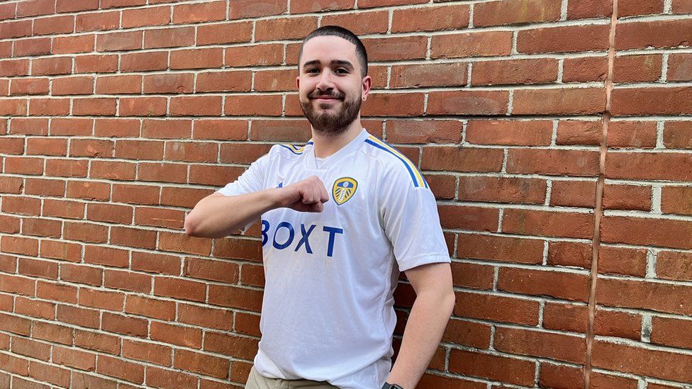 Ollie Ward, in a white Leeds United shirt, in front of a brick wall