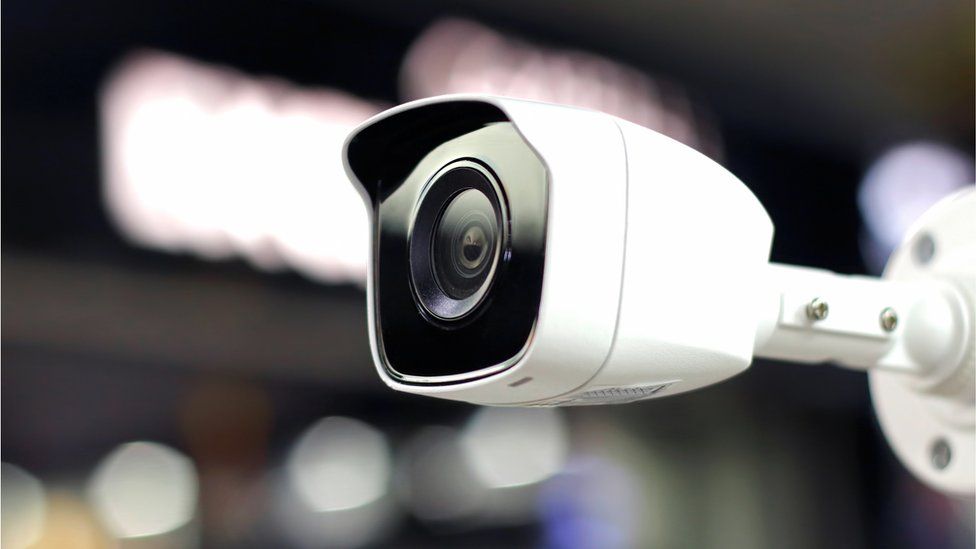 A standard CCTV camera is seen wall-mounted against a blurred background of lights