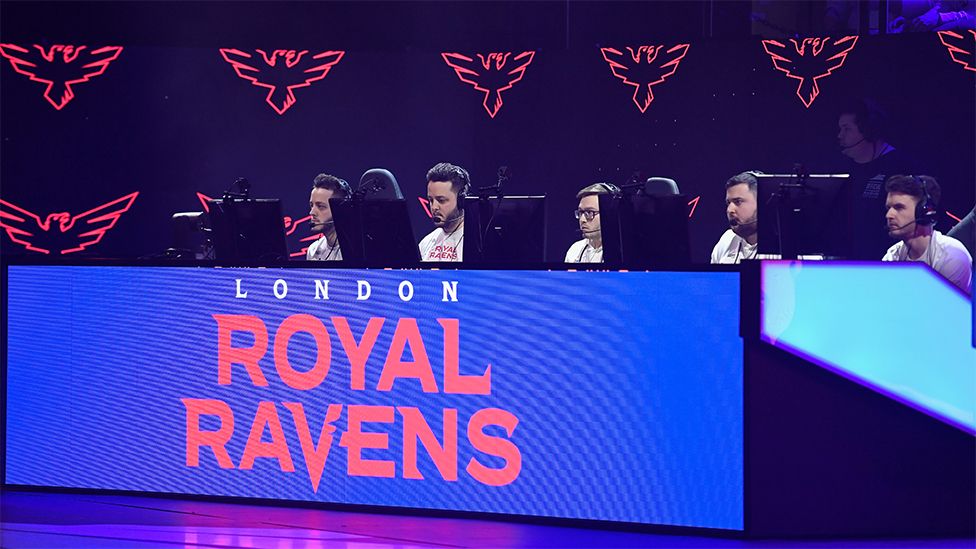 The London Royal Ravens competing in 2020, the competitors are sat behind a blue desk which has the London Royal Ravens logo in white and red writing on a blue background, and they are looking at computer screens