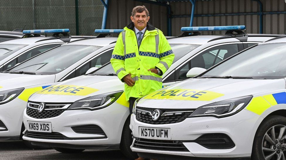 Police & Crime Commissioner, Philip WIlkinson, standing in a high-vis coat with his hands in his pockets beside four police cars.