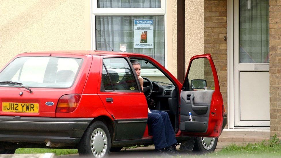Ian Huntley sitting in a car outside his house which has a picture of Holly Wells and Jessica Chapman in the window