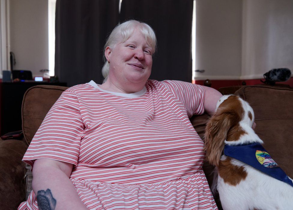 Kayleigh at home on the sofa with dog Rosie next to her. Kayleigh has blond hair and is wearing a pink striped top. She is smiling towards the camera