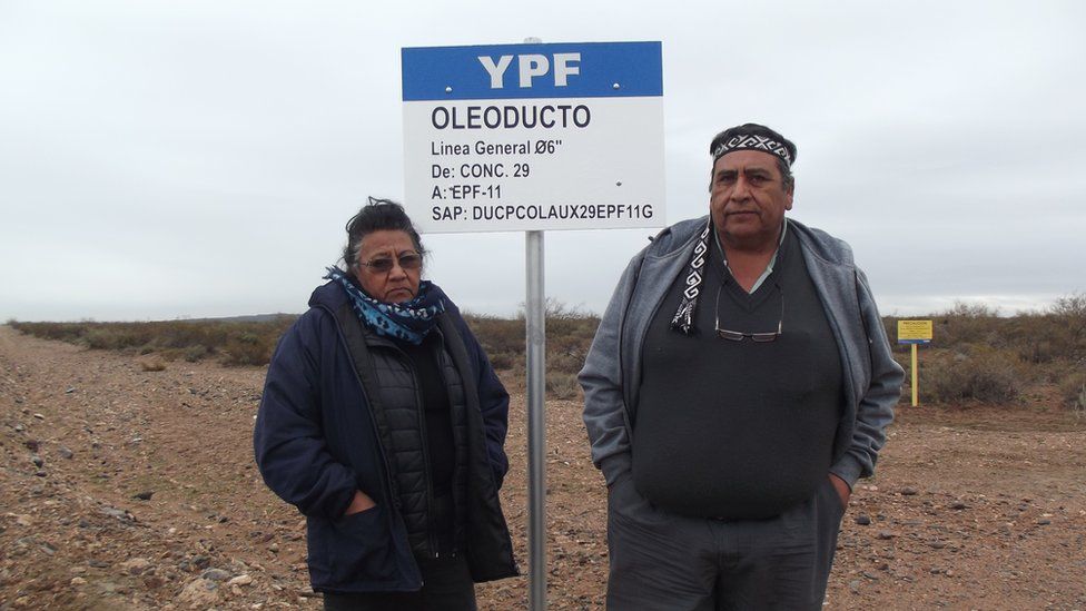Josifa and Albino Campo stand in front of a sign for a YPF oil pipeline