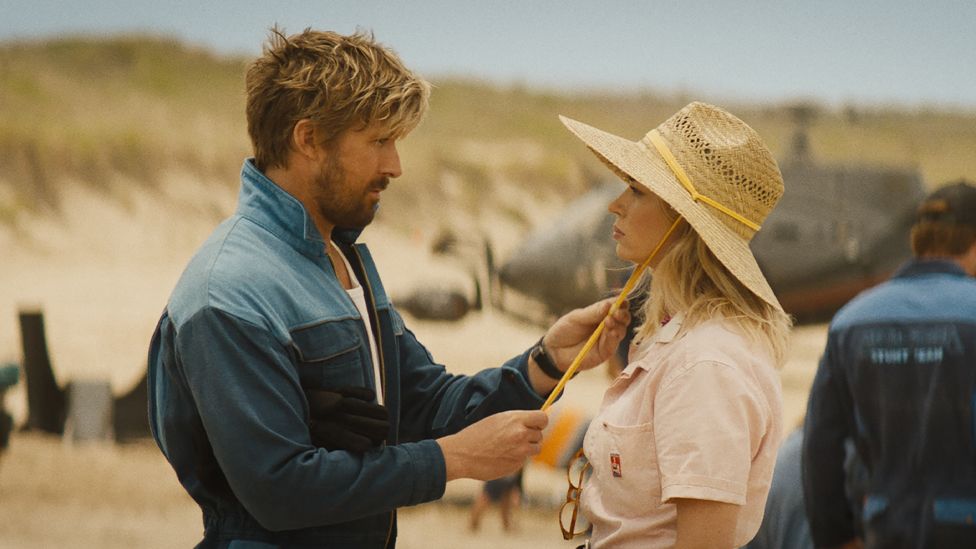 Ryan Gosling is Colt Seavers and Emily Blunt is Judy Moreno in The Fall Guy. Ryan is a white man in his 40s with sandy blonde hair and a short beard. He wears blue overalls, unzipped to reveal a white top. He's facing Emily blunt and holding a yellow ribbon securing her straw hat to her head. Emily is a blonde white woman in her 40s and wears a dusty pink shirt, her glasses looped into a button hole. The pair are pictured outside in a desert scene where sand dunes and a helicopter can be seen in the background.