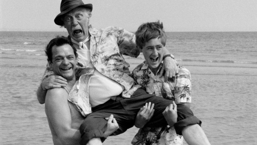 Black and white photo of David Jason (left) and Nicholas Lyndhurst laughing while holding Lennard Pearce over the water at a beach