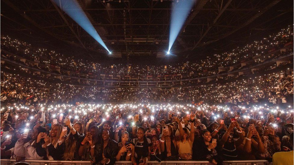 A large crowd in an arena with their phone lights on watching Wizkid perform.