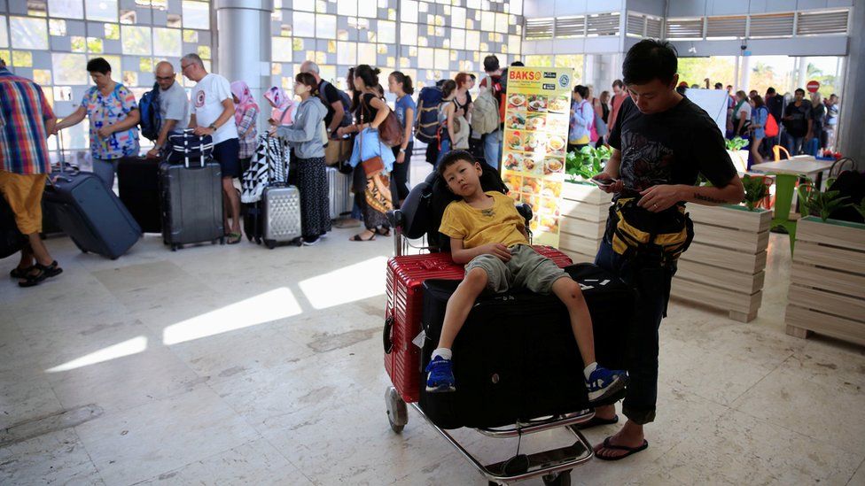 A child sits on top of luggage on a trolley as tourists queue to leave Lombok Island