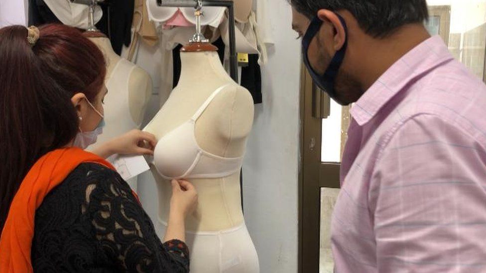 A woman sews a bra together while watched by a man