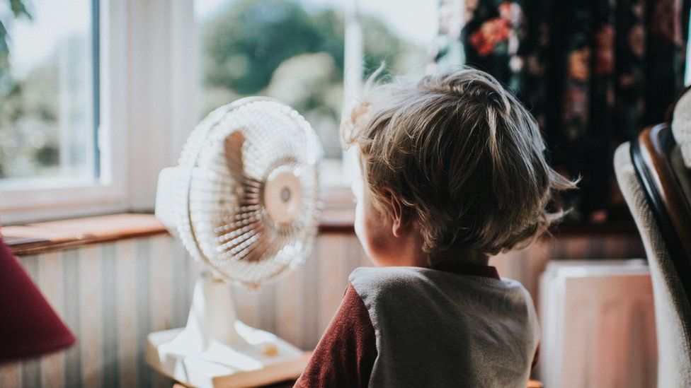 A child looking out a window on a sunny day, with a fan visible in front of them