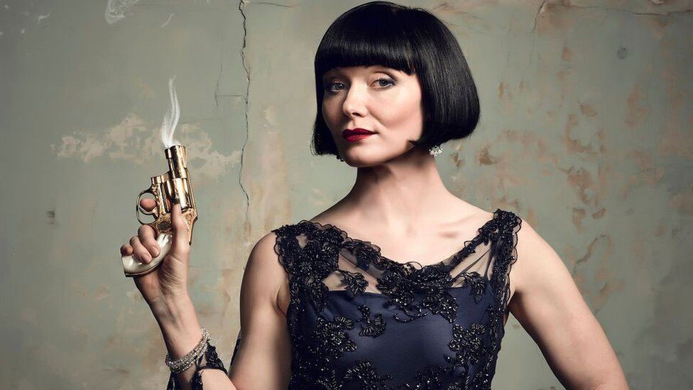 A promotional photo of actress Essie Davis, in character as Miss Fisher, holding a smoking gun