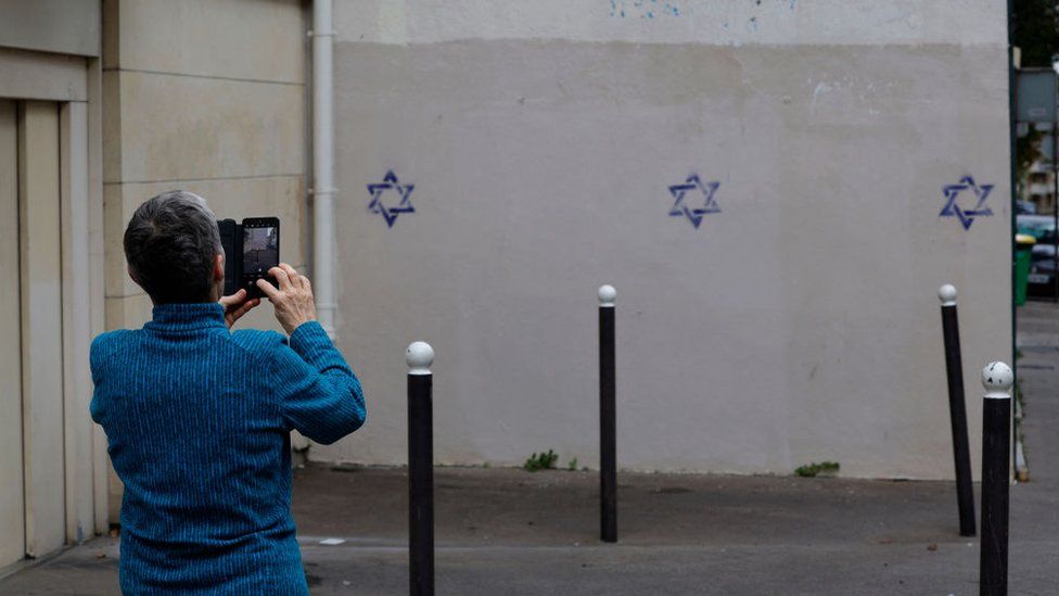 A person photographs a building wall covered with Stars of David