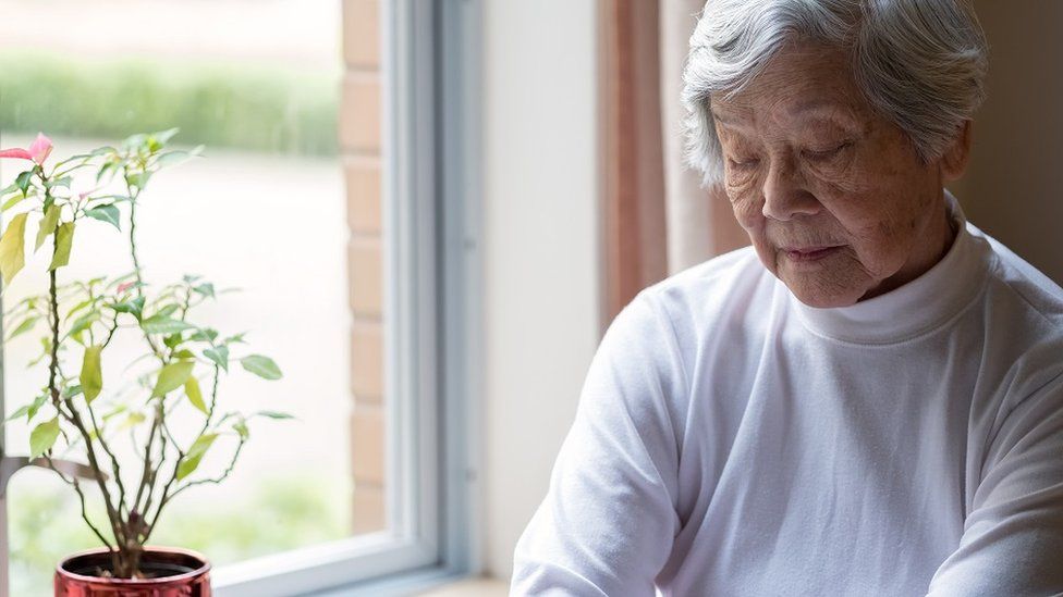 Una mujer de edad, sentada cerca de una ventana con los ojos cerrados