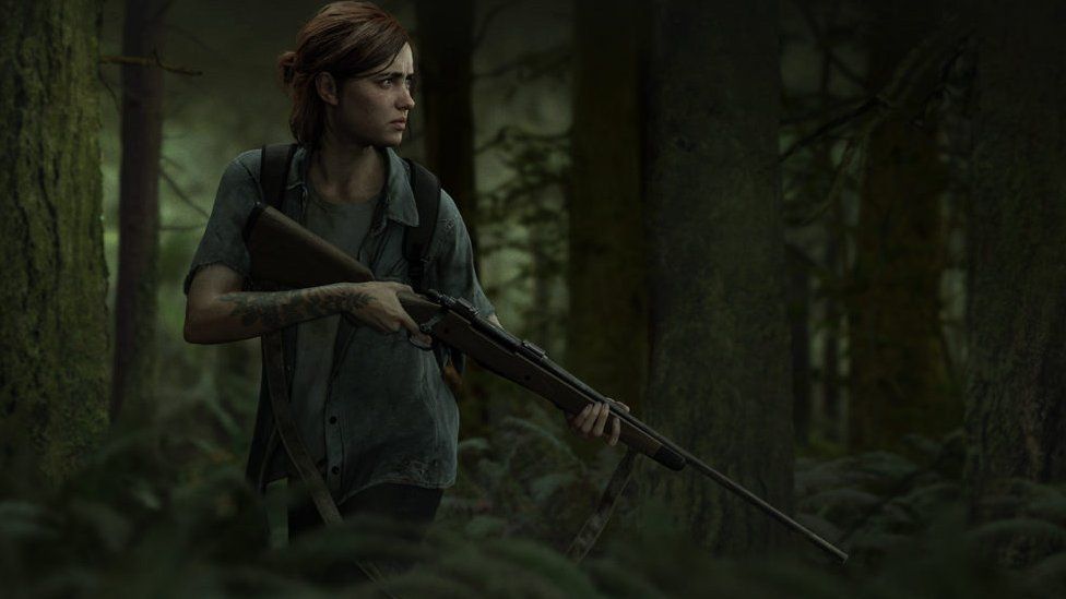 Ellie stands with a gun, looking at something off camera in a green forest