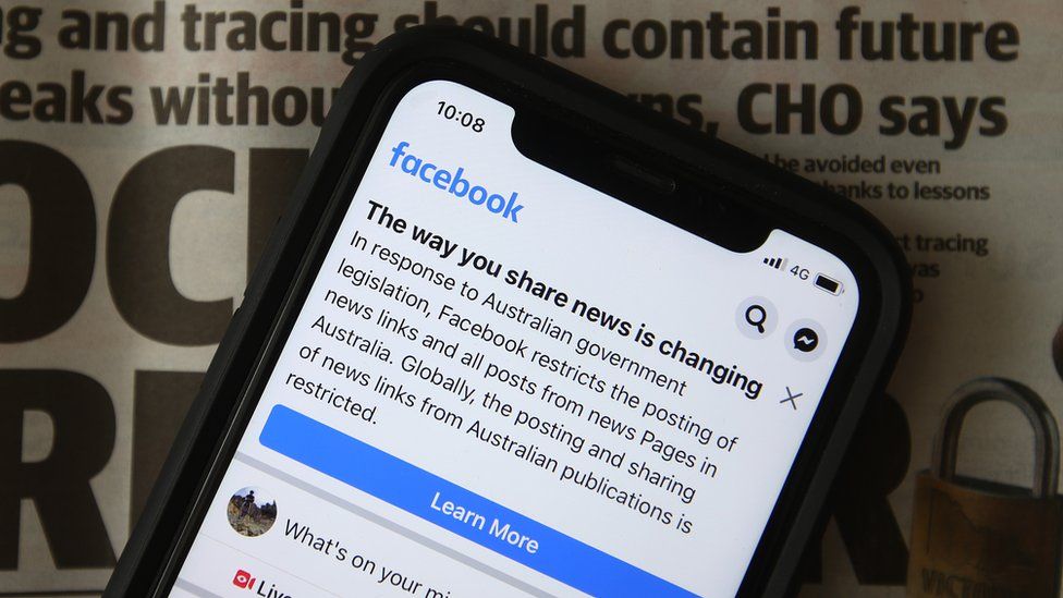 Mobile-phone screen shows message from Facebook announcing its new rules for news sites on its platform. Newspaper underneath the phone.