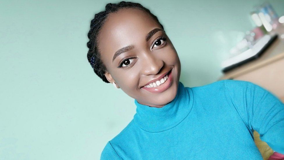 A woman smiles as she pulls a selfie-style pose. She's got her hair tied back and she's wearing a teal-coloured polo neck top. Behind her is a light green wall and a chest of drawers -out of focus but it appears to have containers of beauty products on top of it.