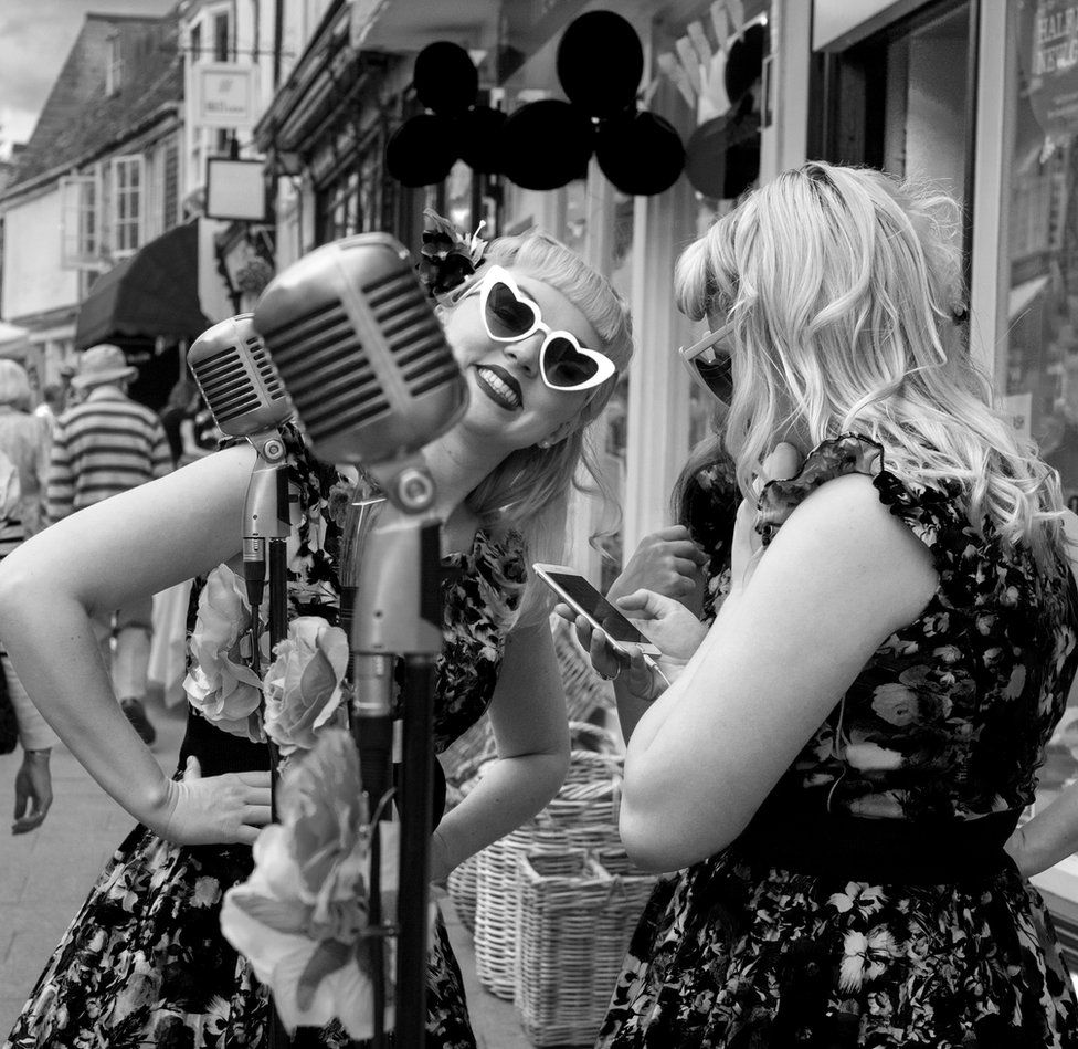 Two 1940s style singers are dressed in similar outfits. one is looking at her phone and the other at the camera