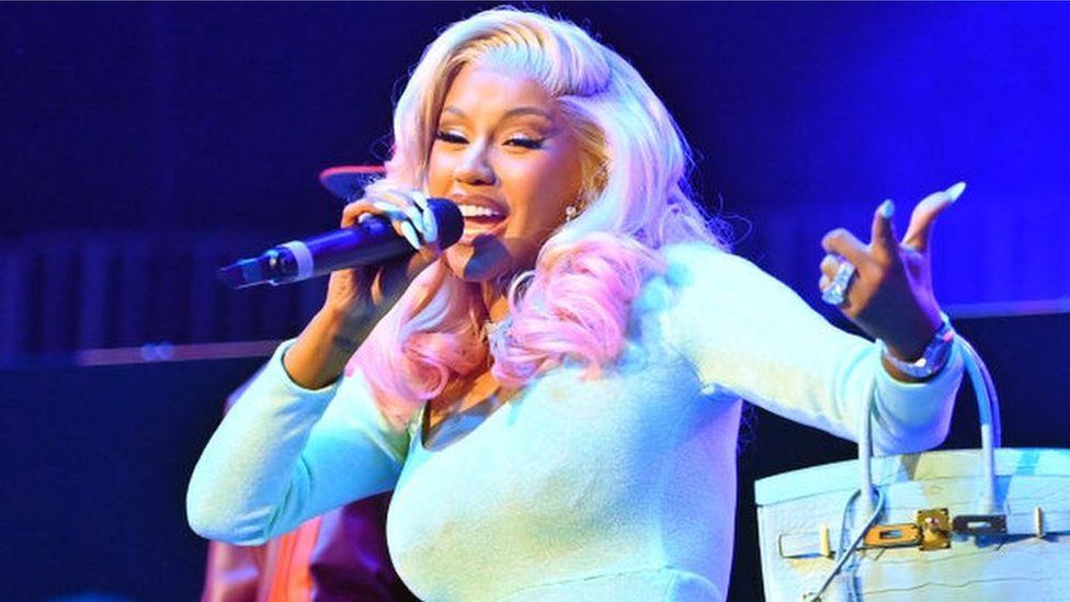 Rapper Cardi B performing on stage. She has bleach blonde hair with is dyed pink at the ends where it has been curled. She is wearing a turquoise long-sleeved dress and is holding a microphone to her mouth with her right hand while she sings/raps. She also has a turquoise handbag hanging from her left arm with the straps around her wrist and her nails are painted turquoise to match her dress.