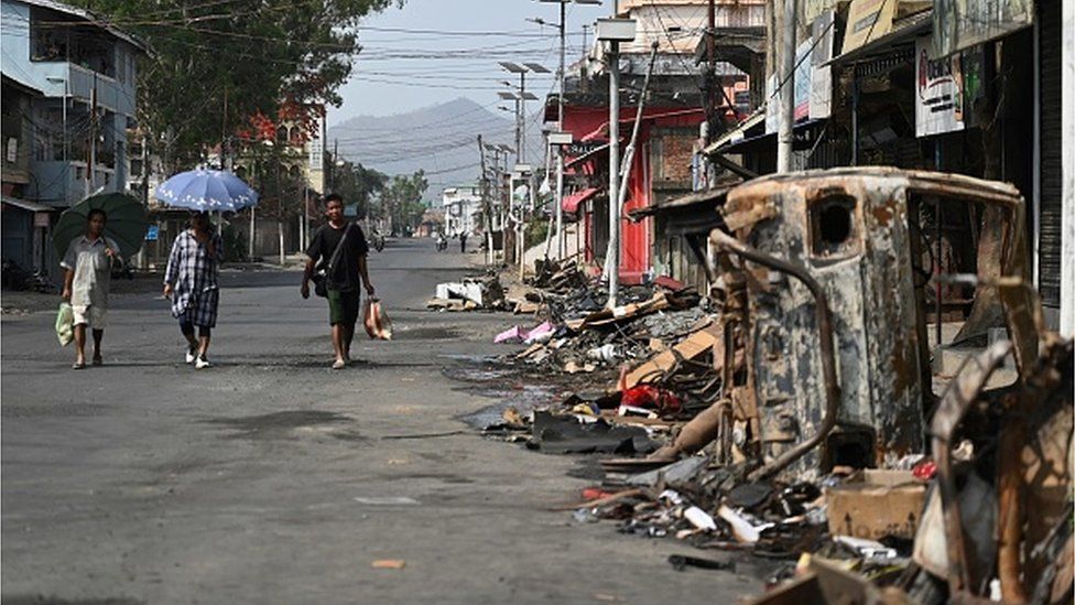 People walk past a burnt vehicle and rubble on a street in Churachandpur in violence hit areas of north-eastern Indian state of Manipur on May 9, 2023.