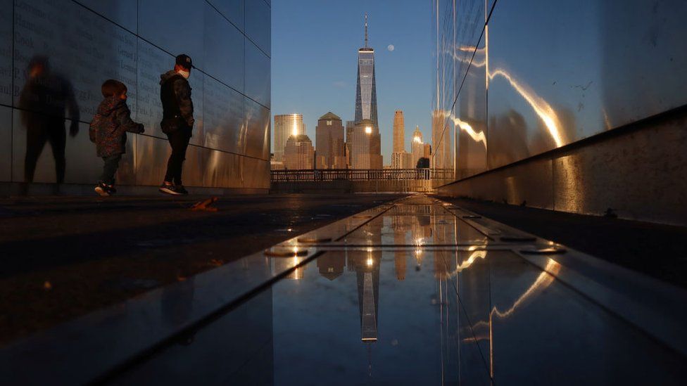 People walk through the Empty Sky 9/11 Memorial as the moon rises next to One World Trade Center in New York City at sunset on February 25, 2021 as seen from Jersey City, New Jersey