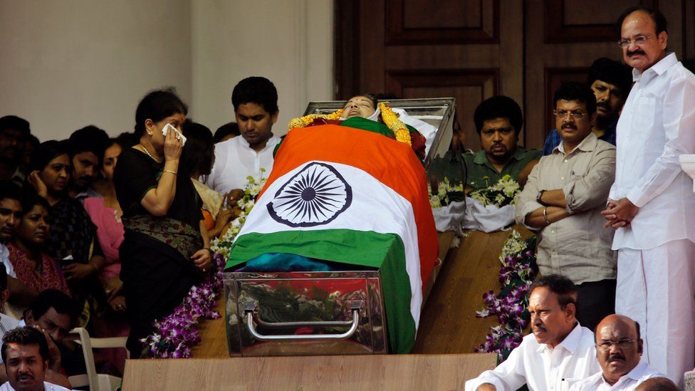 Sasikala Natarajan, left standing, a close friend of India"s Tamil Nadu state former Chief Minister Jayaram Jayalalithaa, wipes her tears next to Jayalalithaa"s body wrapped in the national flag and kept for public viewing outside an auditorium in Chennai, India